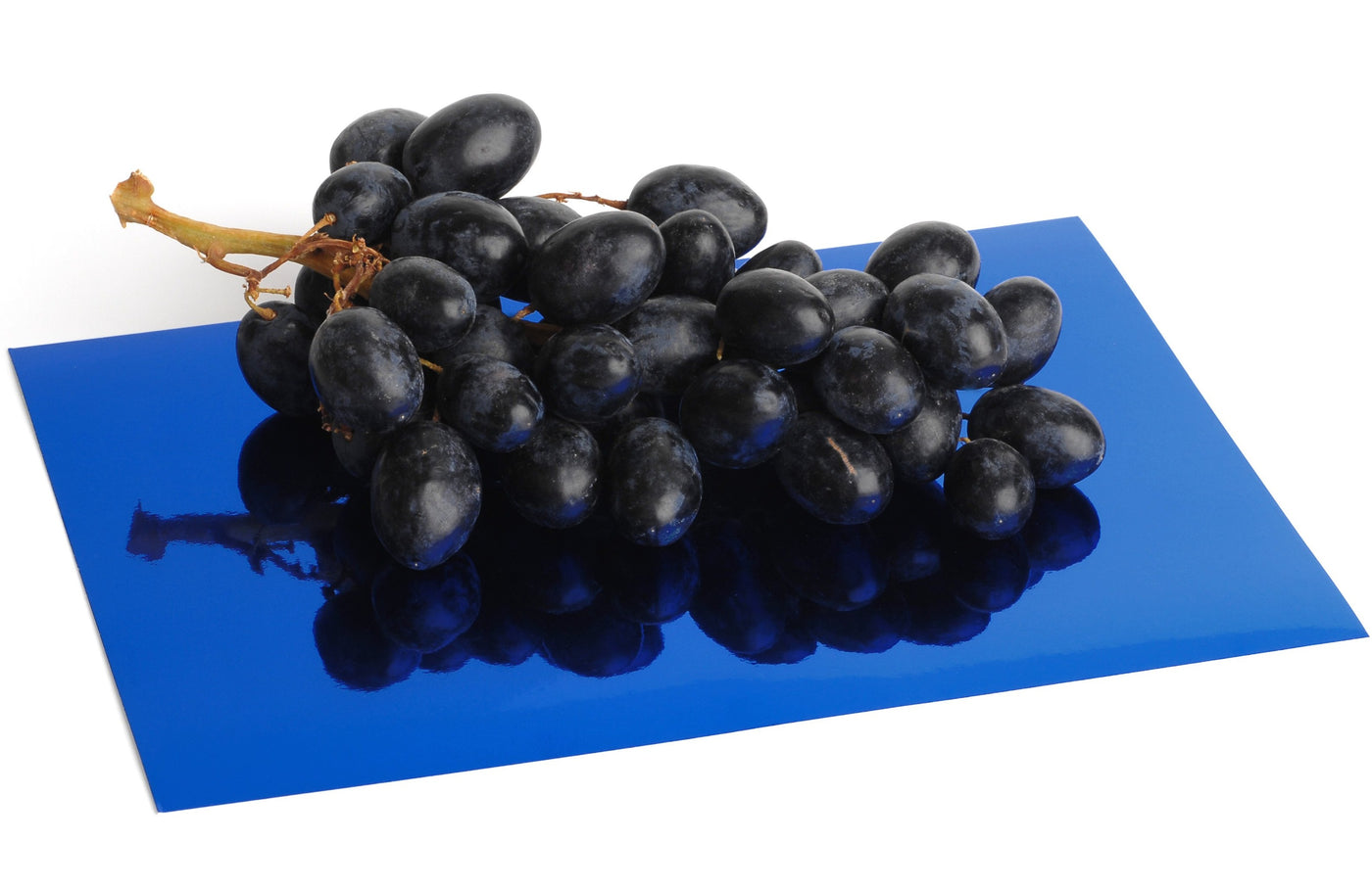 A sheet of deep blue mirrored foil board. Highly reflective with an intense blue color. A bunch of grapes sit atop the mirror foil board and are reflected with near perfect fidelity.